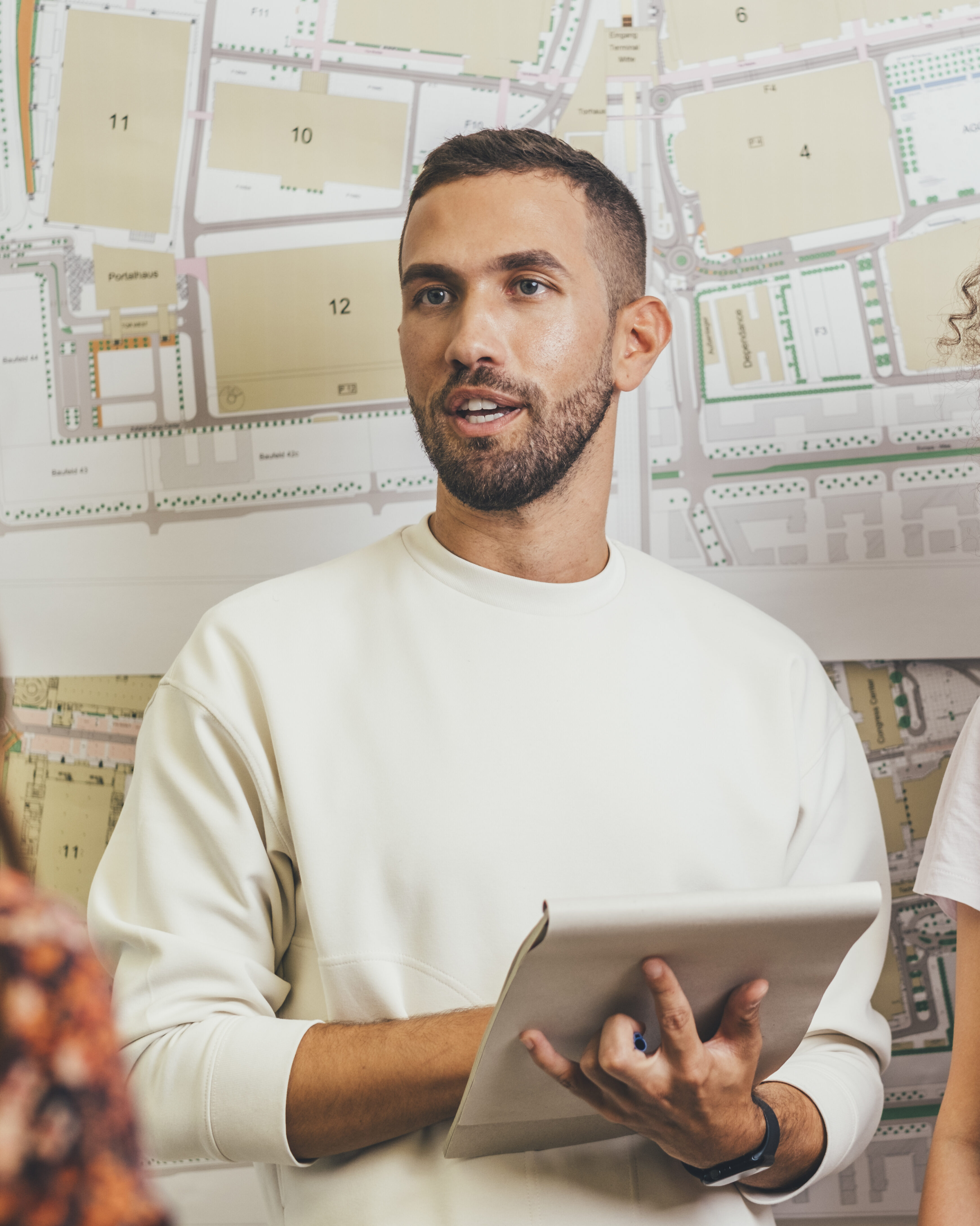 Ein Mann steht vor einer Notizwand, hält einen Block in der Hand und trägt etwas vor oder unterhält sich mit weiteren Personen.