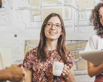 Eine Frau steht mit weiteren Personen vor einer Notizwand und unterhält sich. Sie hält eine Tasse in der Hand.