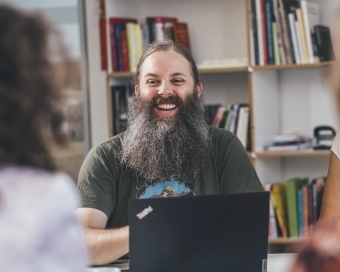Mehrere Personen sitzen an einem Tisch und unterhalten sich. Der Kamerafokus liegt auf einem Mann und seinem Laptop.